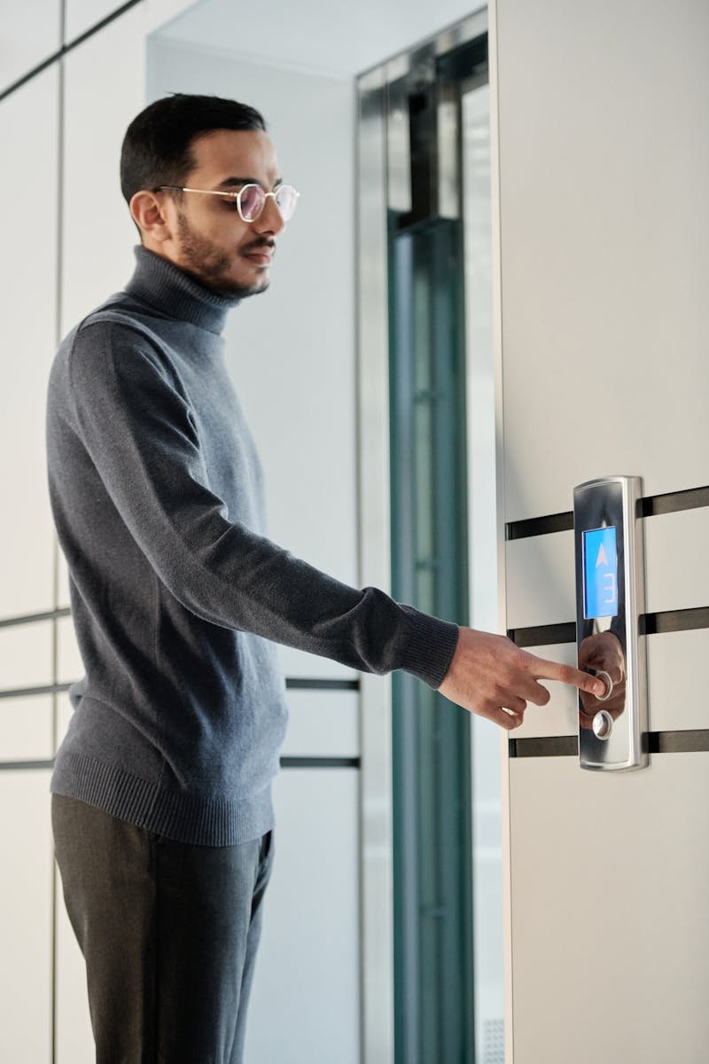 A man wearing glasses and a turtleneck presses an elevator button in a sleek, modern building interior.